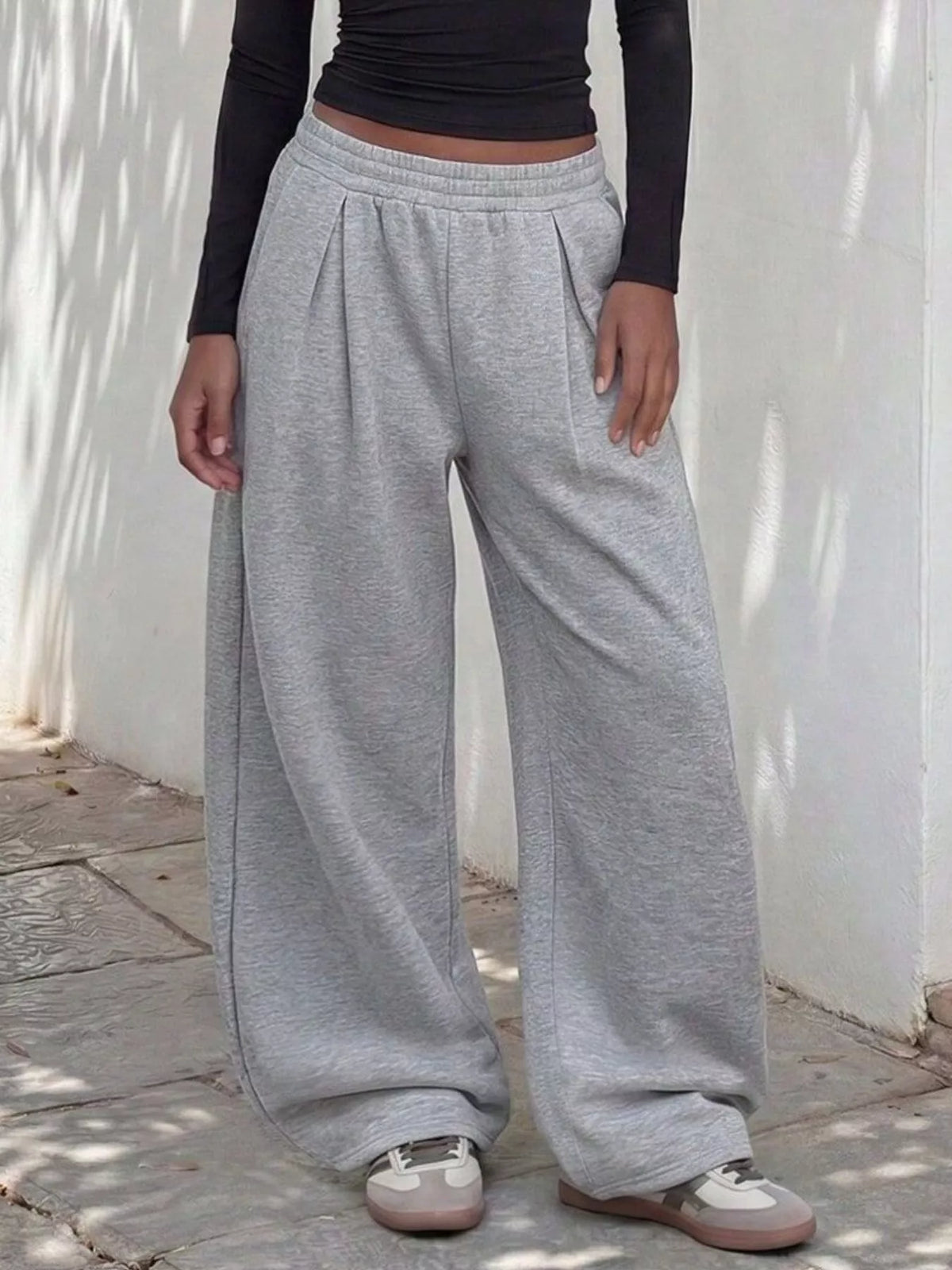 Model wearing gray wide-leg sweatpants with an elastic waistband, front pleats, and sneakers beside a white wall.